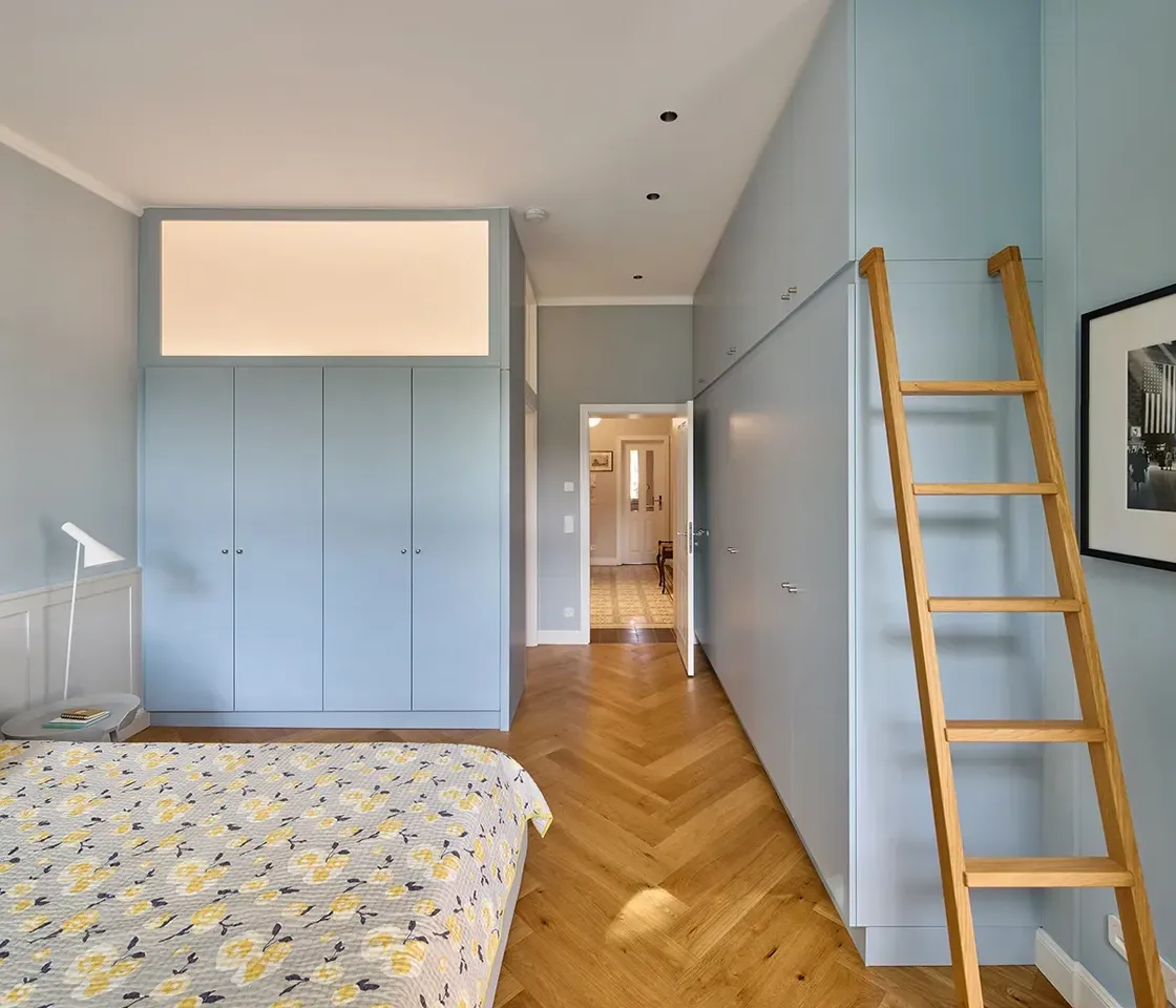 Schlafzimmer einer Maisonettewohnung mit maßgefertigten Einbauten in Hellblau, Leiter zum oberen Stauraum und Eichenparkett im Fischgrätmuster.